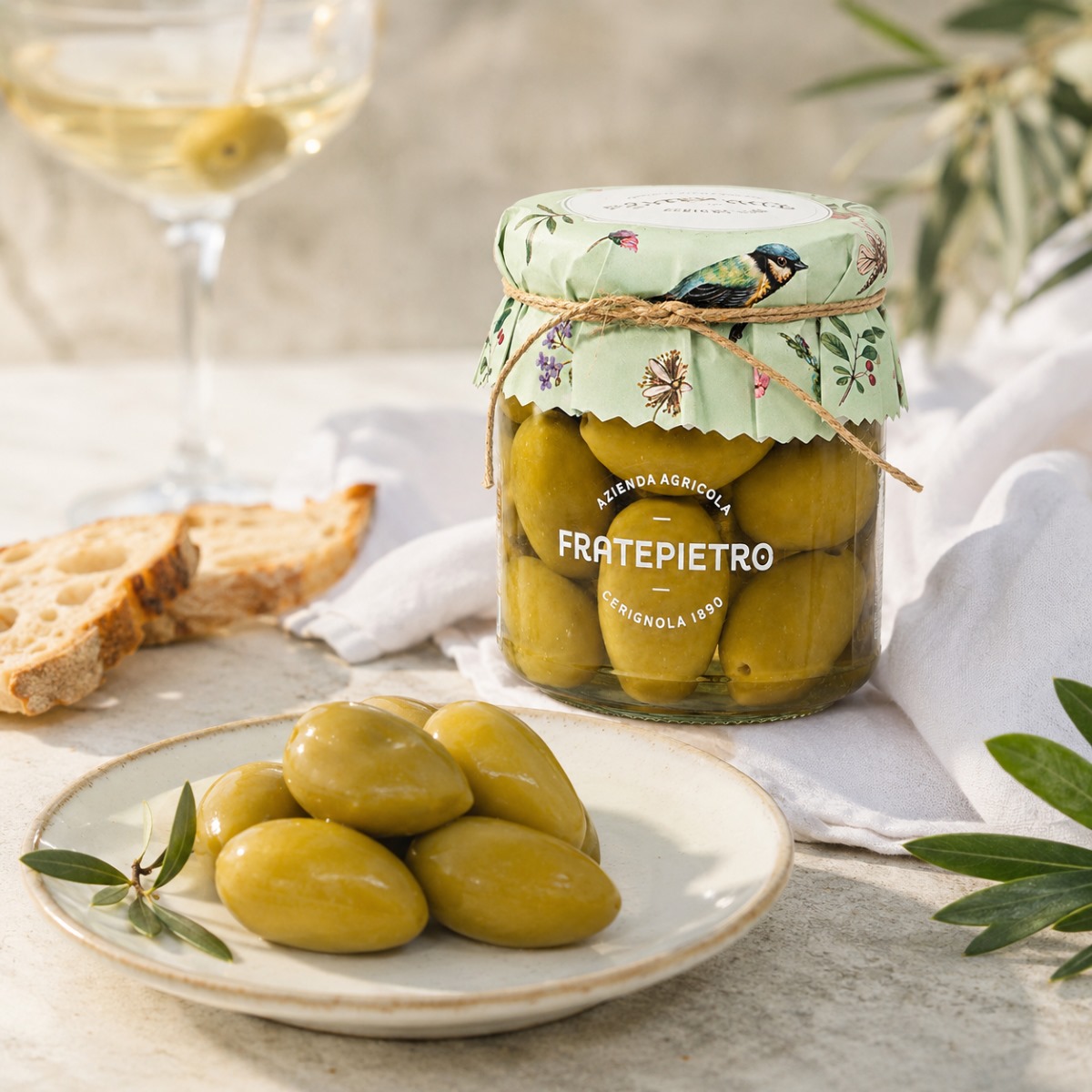 Aceitunas Verdes Bella di Cerignola Fratepietro Servidas Aceitunas Verdes Bella di Cerignola Fratepietro servidas con pan, copa y hojas de olivo en escena mediterránea