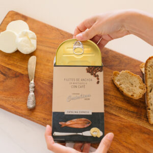 Manos abriendo lata de filetes de anchoa en mantequilla con café Catalina Expreso sobre tabla de madera con pan y queso.
