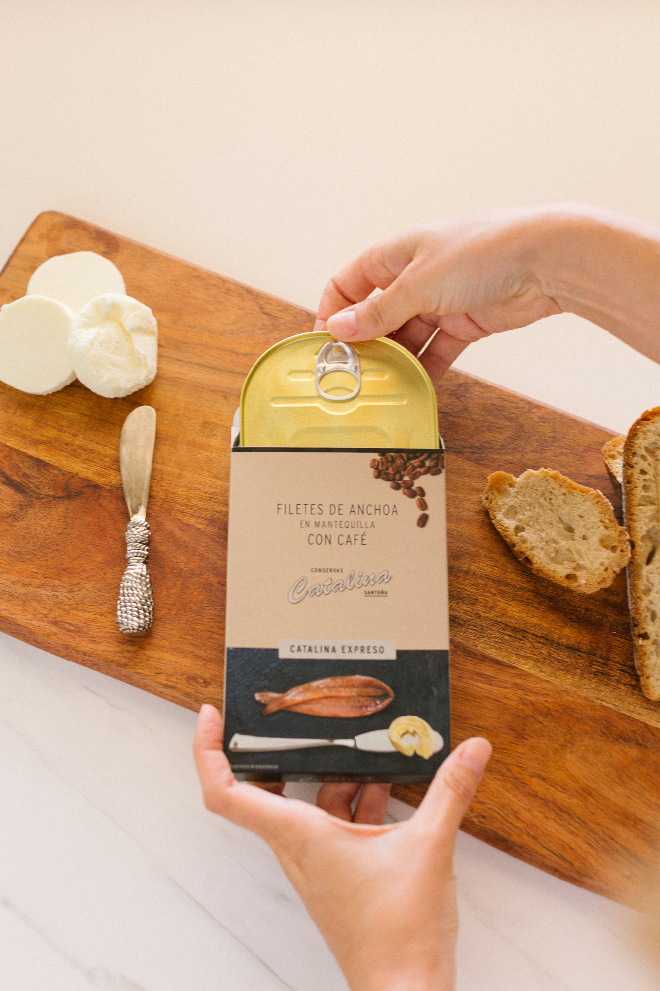 Filetes de Anchoa en Mantequilla con Café Catalina – Presentación Gourmet Manos abriendo lata de filetes de anchoa en mantequilla con café Catalina Expreso sobre tabla de madera con pan y queso.